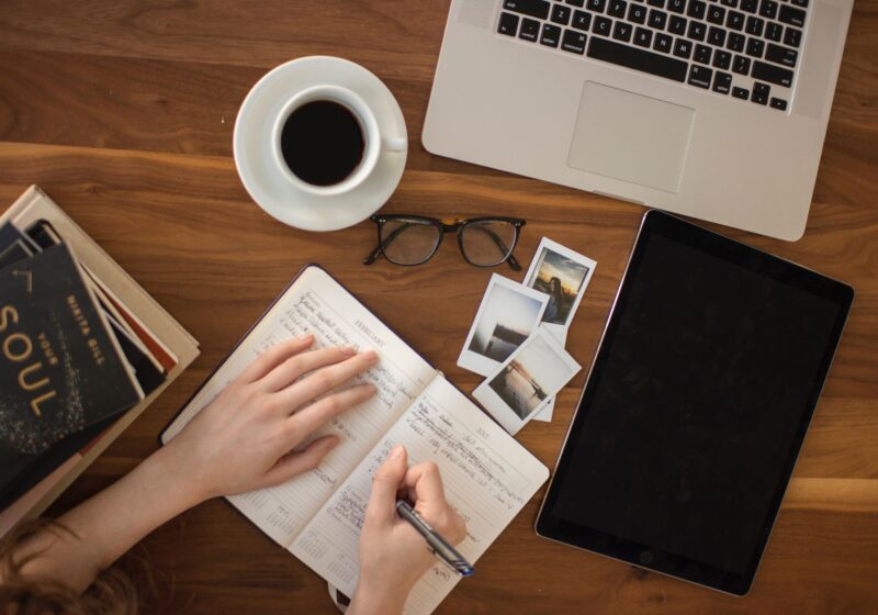 a table with a laptop, coffee, and a girl writing in a journal