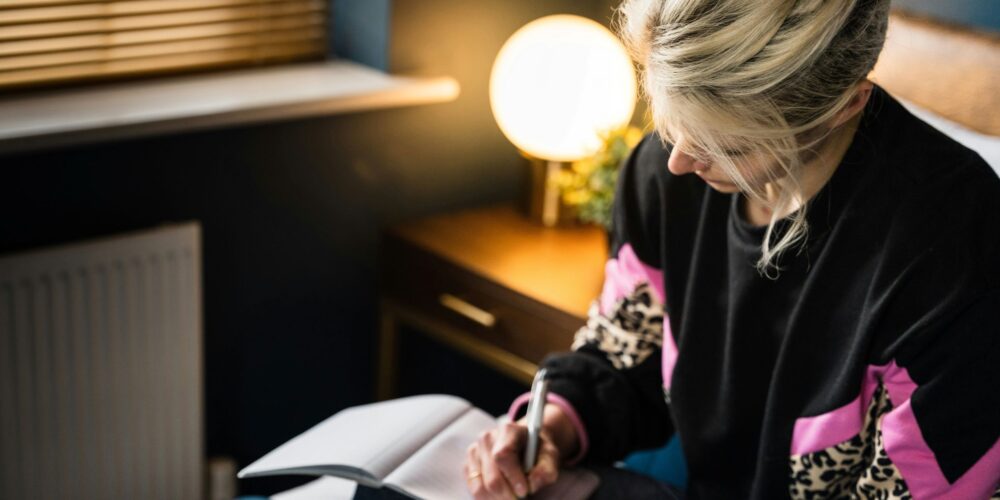 A young woman sitting on a bed, peacefully writing in her journal using prompts for self-care and mental health, creating a calm and reflective atmosphere.