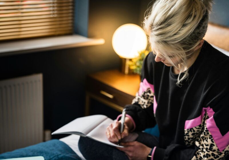 A young woman sitting on a bed, peacefully writing in her journal using prompts for self-care and mental health, creating a calm and reflective atmosphere.