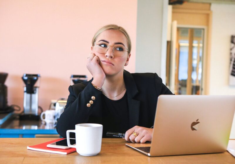 A thoughtful woman sits at her work desk with a coffee mug and laptop, representing the mental load and daily reflections of a 9-5 job routine.