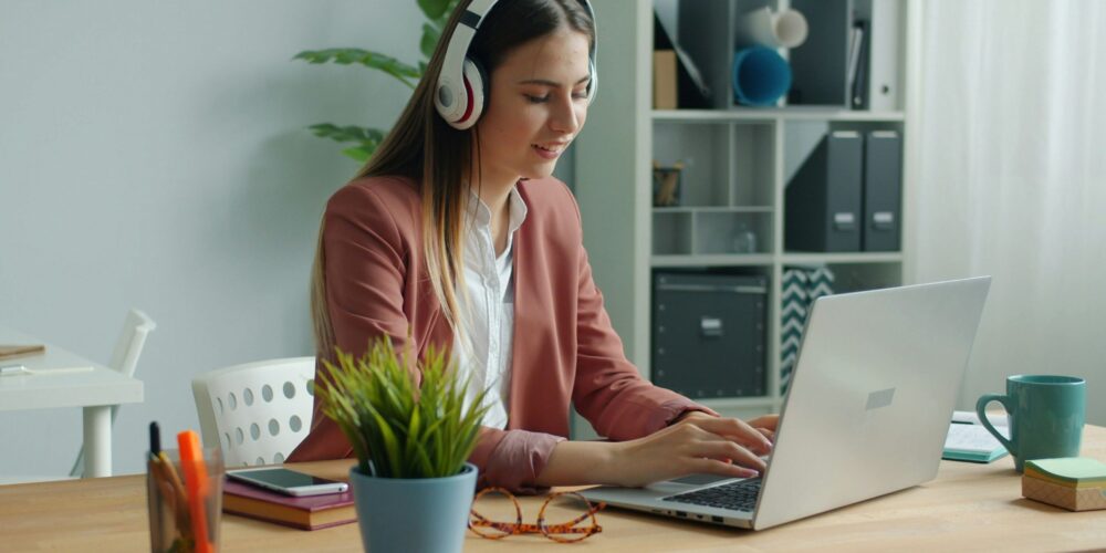 A woman working remotely on a laptop at a tidy desk with a notebook and coffee beside her. She is sitting in a bright, calm room, showing a simple, self-care-friendly work setup that supports focus and better work performance.