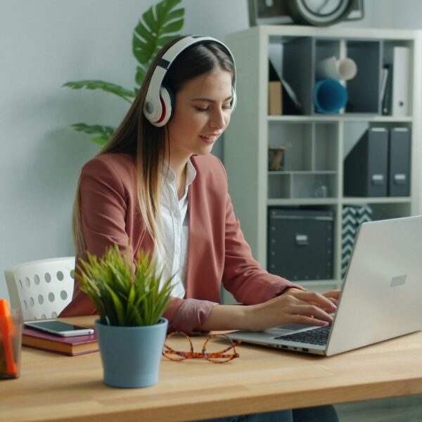 A woman working remotely on a laptop at a tidy desk with a notebook and coffee beside her. She is sitting in a bright, calm room, showing a simple, self-care-friendly work setup that supports focus and better work performance.