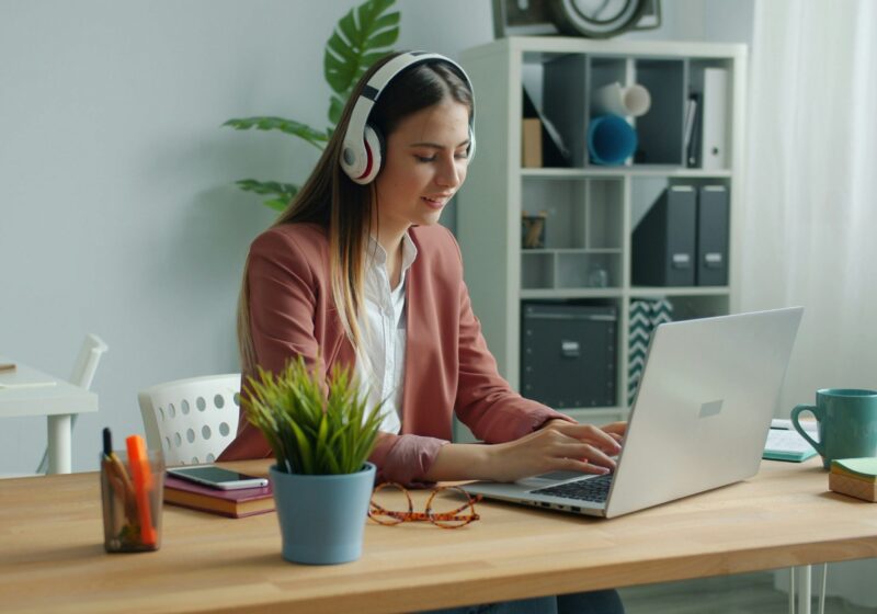 A woman working remotely on a laptop at a tidy desk with a notebook and coffee beside her. She is sitting in a bright, calm room, showing a simple, self-care-friendly work setup that supports focus and better work performance.