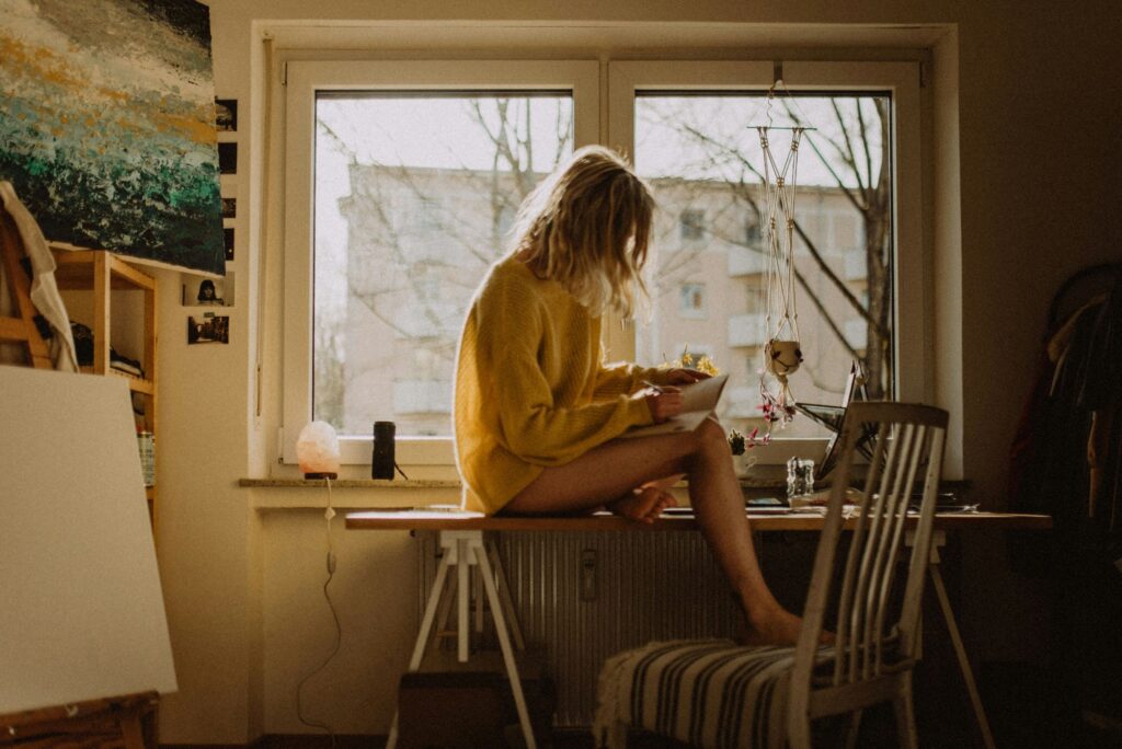 A woman practicing mindful living at home while journaling in a calm, cozy space. She is seated comfortably, fully present in the moment, reflecting with awareness and intention—symbolizing simple tips for a more awakened life through journaling and self-reflection.