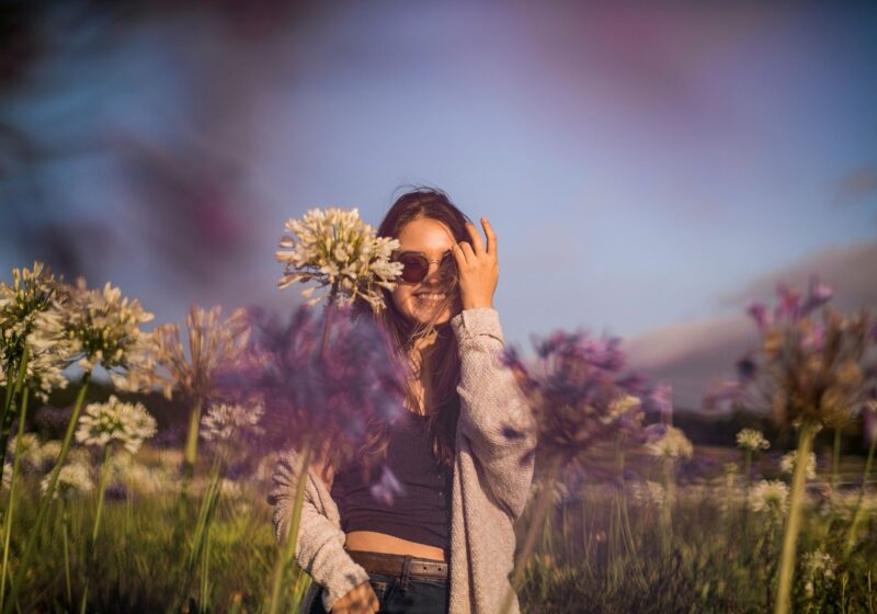 A young woman standing in a blooming spring garden filled with soft pastel flowers, gently touching petals with her fingertips.